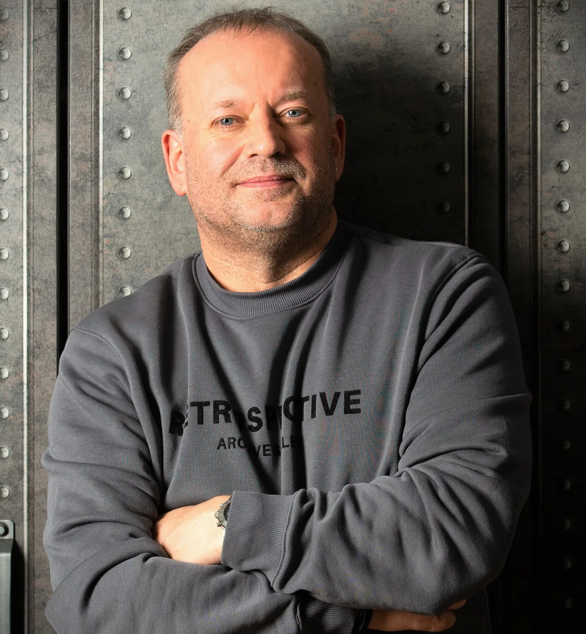 Gerald Stefan Allmann mit verschränkten Armen, graues Sweatshirt vor industrieller Metallwand